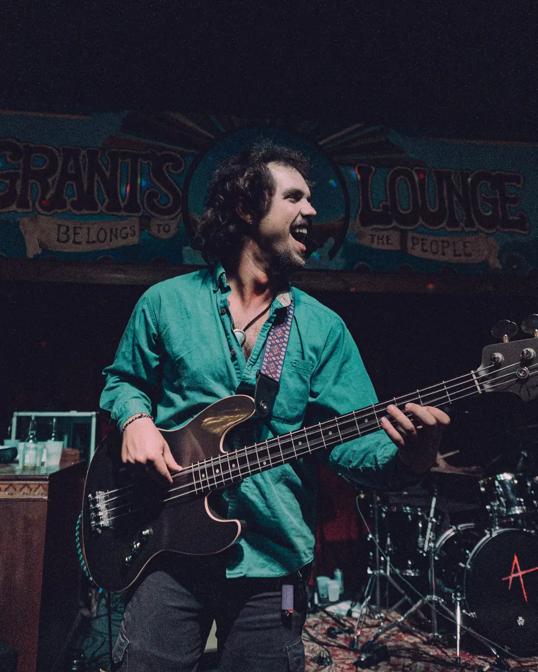 Erik Wolstenholme playing bass in a green shirt under the Grant's Lounge backdrop.